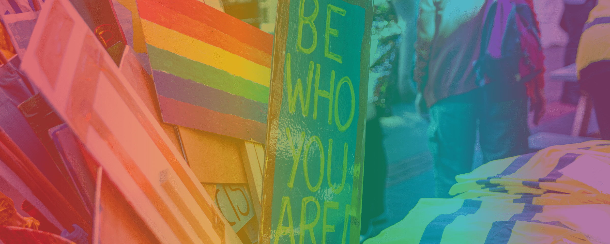 An image with various placards including one with a horizontal rainbow flag and one with text that reads 'BE WHO YOU ARE!