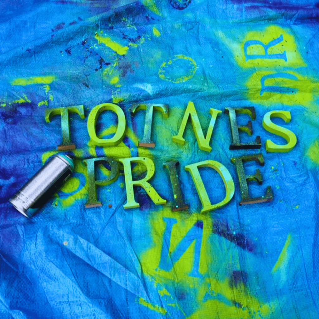  Blue tarpaulin with large cardboard letters spelling out 'Totnes Pride'. On the left of the letters is a spray can laying horizontally. The letters and tarpaulin are randomly sprayed with green and dark blue spray paint.